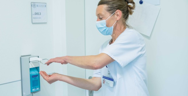Andrea Kollotzke mit Mund-Nasen-Schutz, druckt auf einen Spender für Desinfektionsmittel. Mit der rechten Hand fängt sie die Flüssigkeit auf