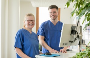 Zwei Pflegefachpersonen stehen vor einem Computer und lächeln in die Kamera